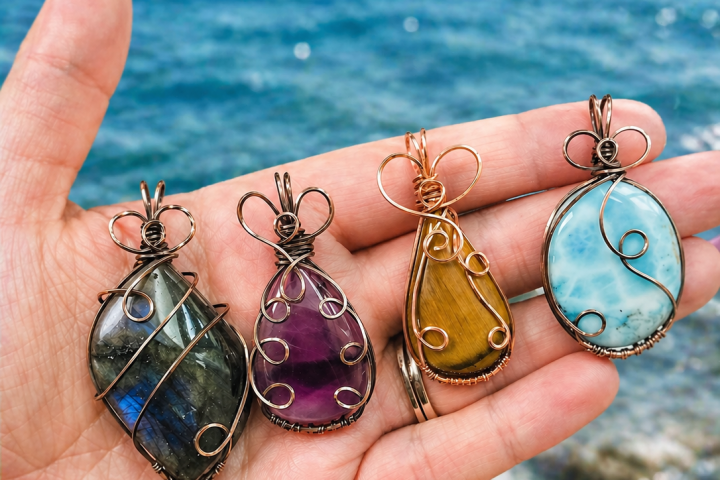 Hand holding four wire-wrapped crystal pendants by the sea.