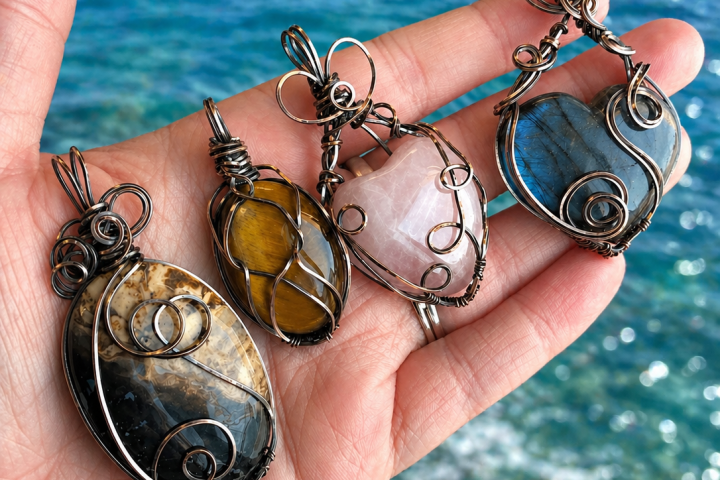 Four wire-wrapped stone pendants on a hand by the sea.