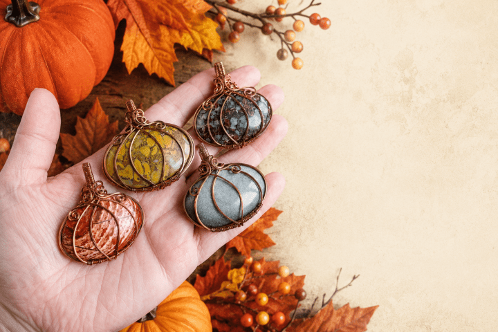Hand holding four stones wrapped in wire, surrounded by autumn leaves and pumpkins.