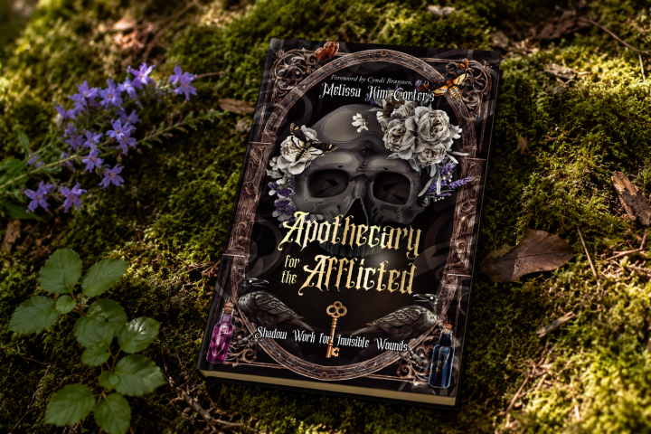 Book titled 'Apothecary for the Afflicted' on mossy ground with flowers and leaves.