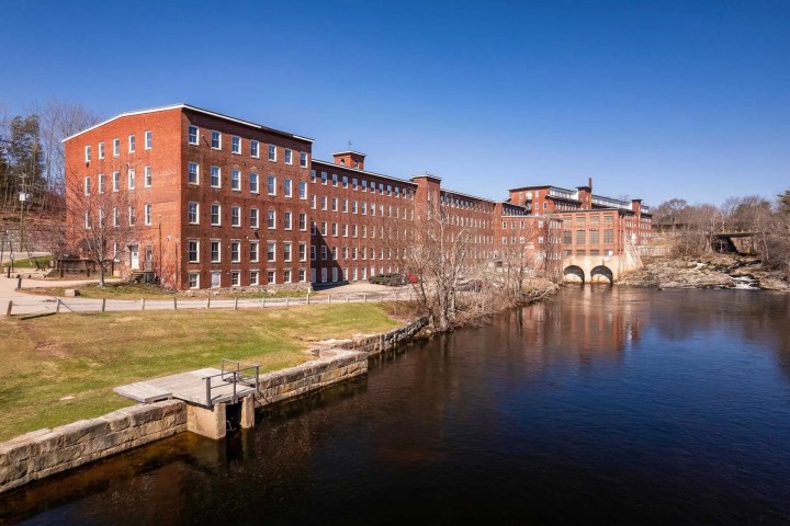 Historic red brick mill building by a river under a clear blue sky.