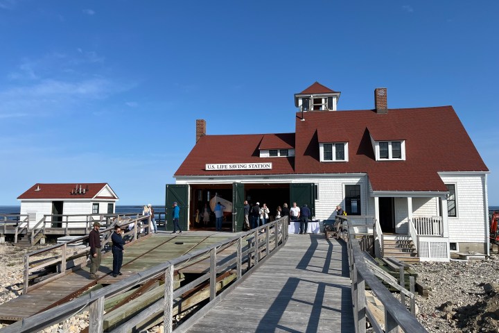 Historic life-saving station with red roof, people touring, and clear blue sky.