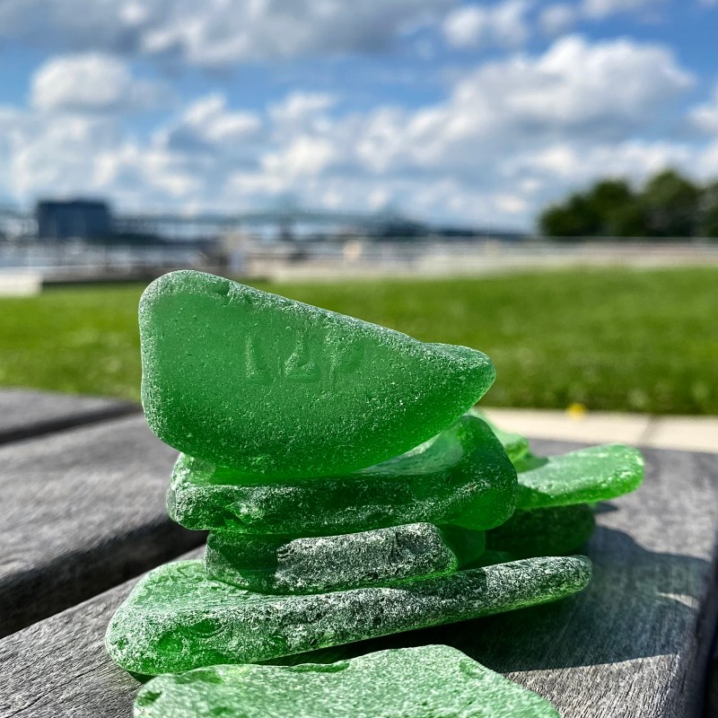 Pile of green sea glass on a wooden bench, with a blurred park and sky in the background.