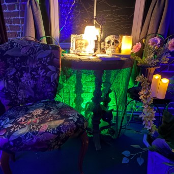 Curved chair, table with candles, skull, flowers, curtains, eerie blue-green lighting.